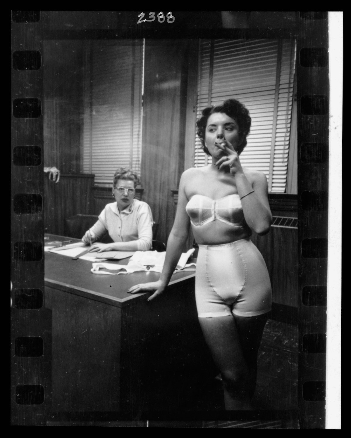 Lingerie model, wearing a girdle and strapless bra, smoking in an office; in the background a woman sits at a desk